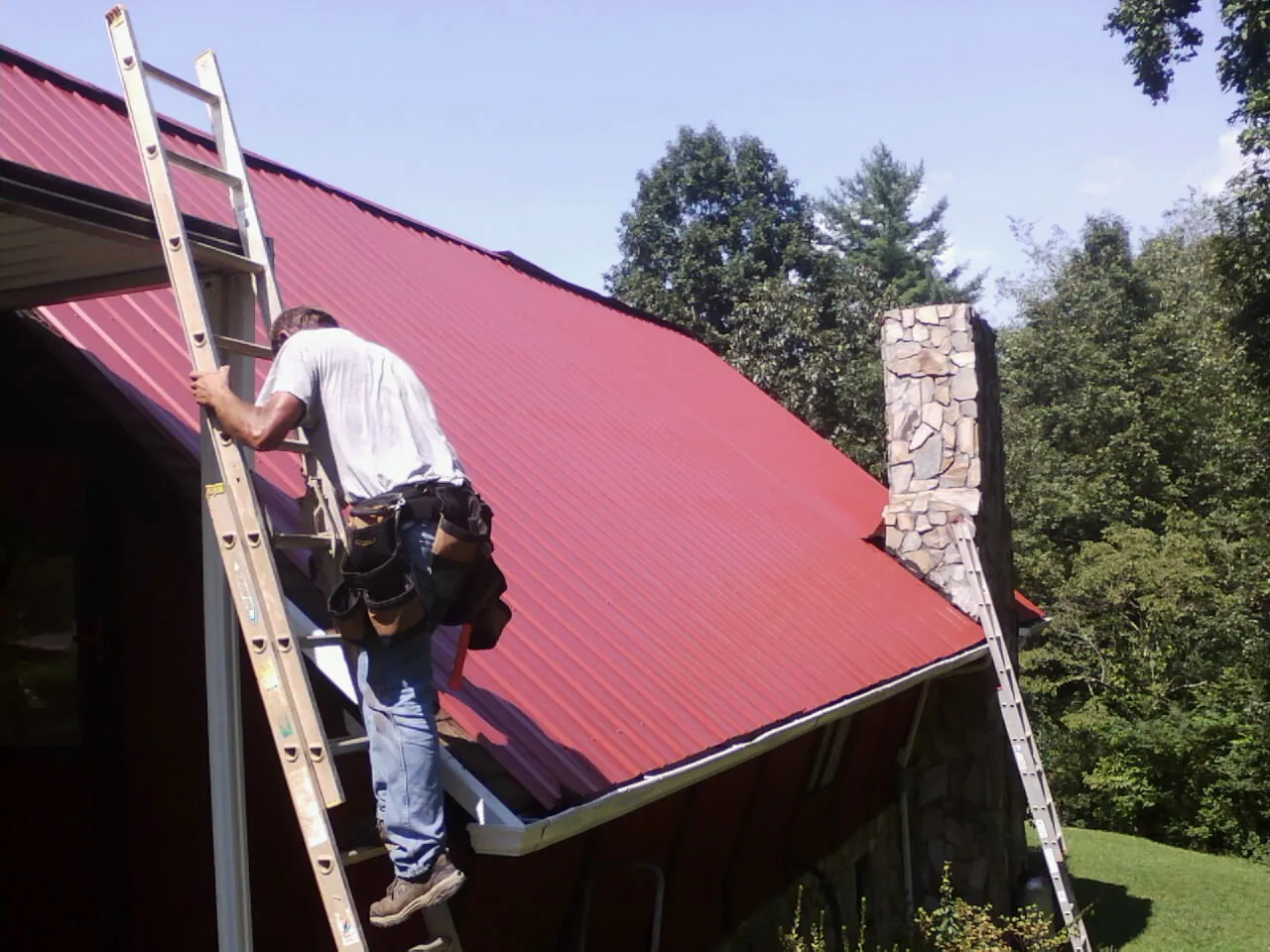 Roofer on a ladder beside a red metal roof and stone chimney — Soffit & Fascia Repair by Roofing Brothers in Yancey